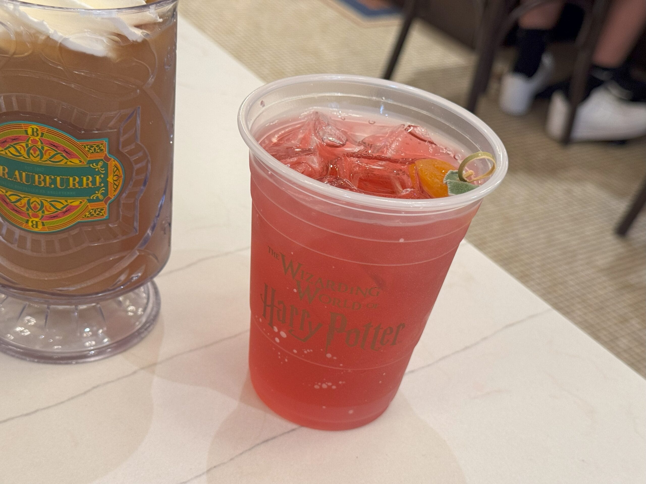 A red iced drink in a "Wizarding World of Harry Potter" cup at Epic Universe, beside a Butterbeer mug.