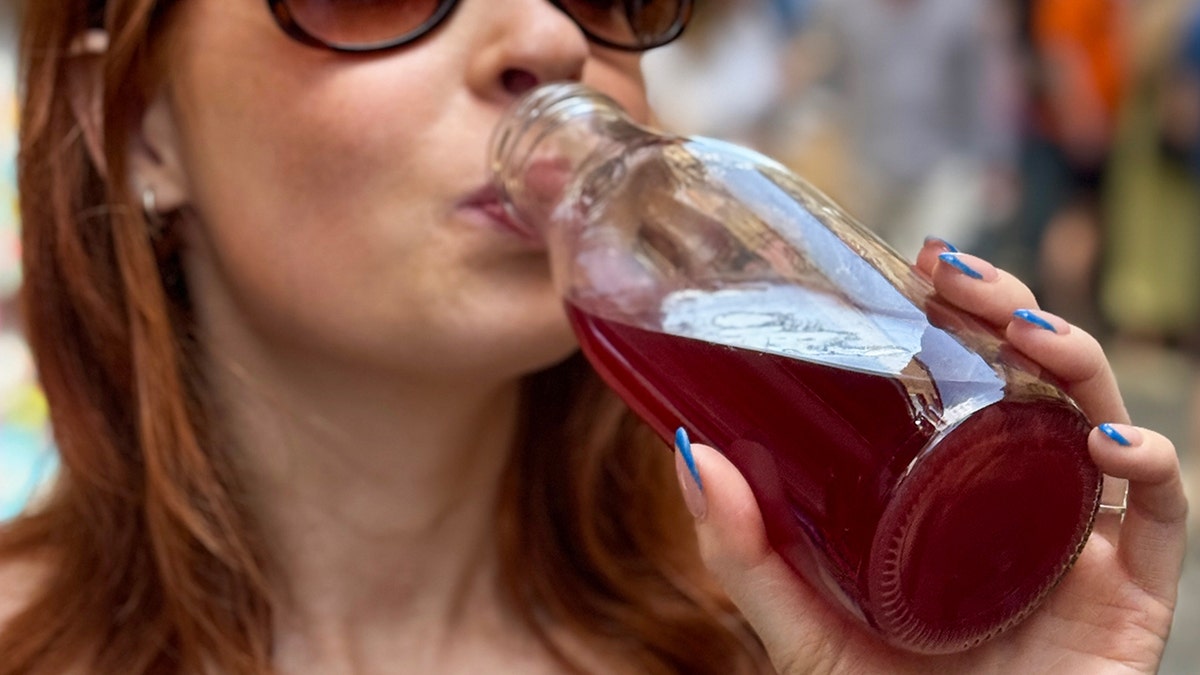 A red-headed woman is drinking kombucha from a glass.