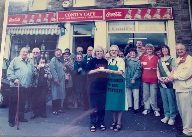 Ida wears an apron which reads "I'm supposed to be retired" as she poses with her business owner daughter and their regular customers