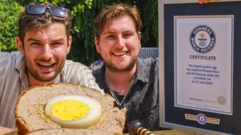 Oli Paterson and Phoenix Ross Two men, both with brown hair and short beards, are seated at an outdoor table holding a Guinness World Records certificate and are smiling at the camera. In front of them lies a massive Scotch egg, sliced in half to reveal the egg inside.