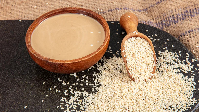 Tahini in a wooden bowl next to a wooden spoon overflowing with sesame seeds on a brown plate