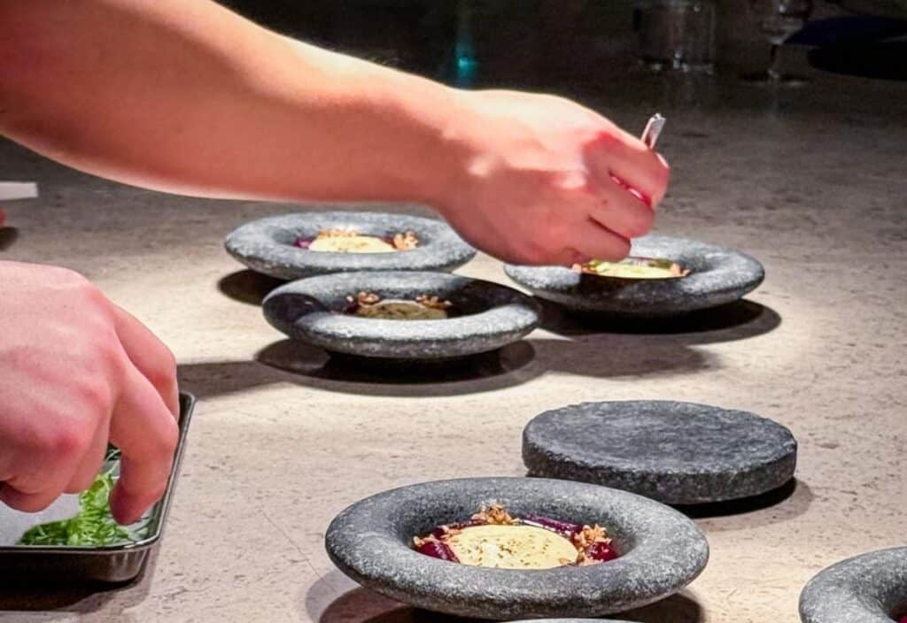 A person uses tweezers to plate food on stone dishes, carefully arranging garnish on several identical plates set on a countertop, showcasing the precision seen at chef’s tables and in tasting menus.