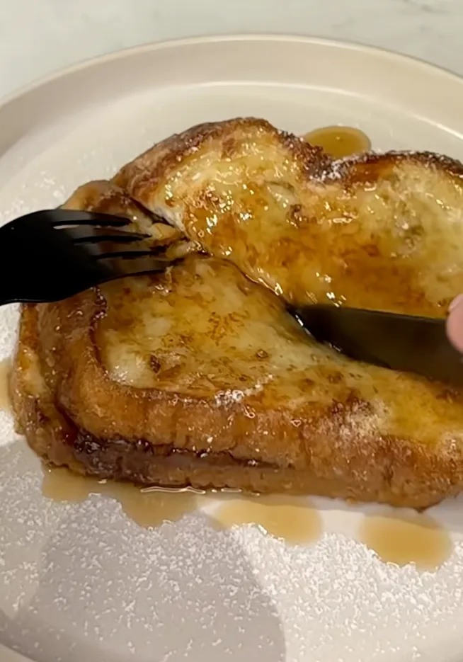 A fork and knife cut into French toast topped with syrup and a dusting of powdered sugar on a plate