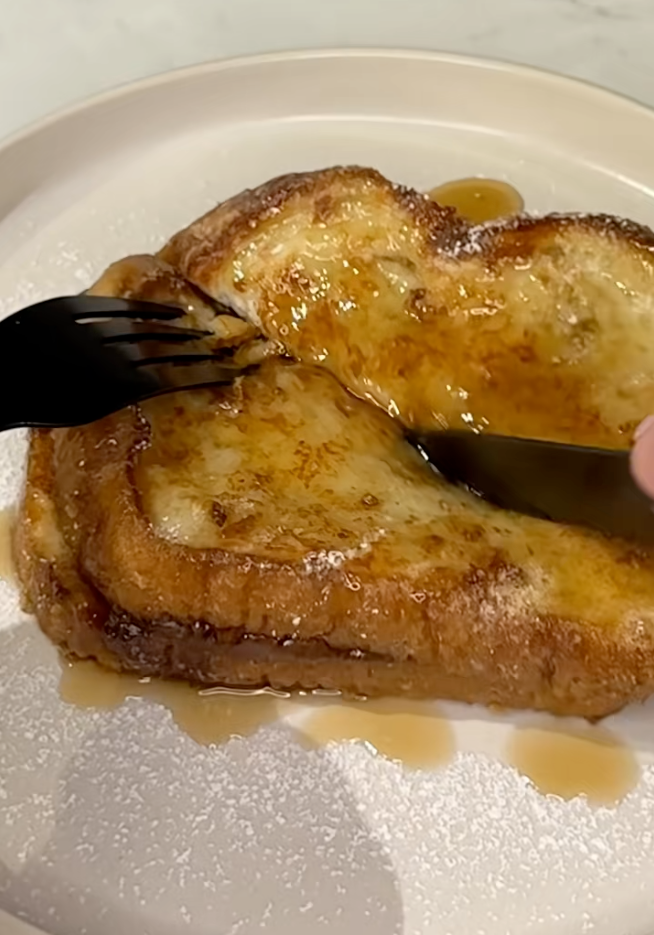 A fork and knife cut into French toast topped with syrup and a dusting of powdered sugar on a plate