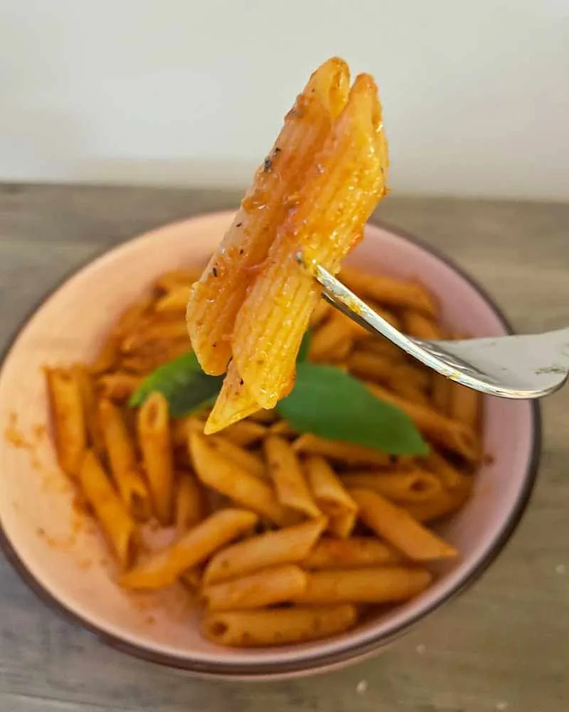 Penne pasta with a red sauce and basil leaf on a fork, served in a pink bowl.