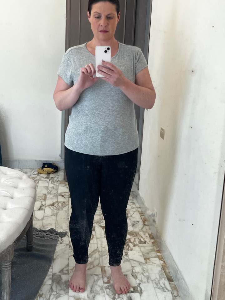 Woman taking a mirror selfie in gray shirt and black pants.