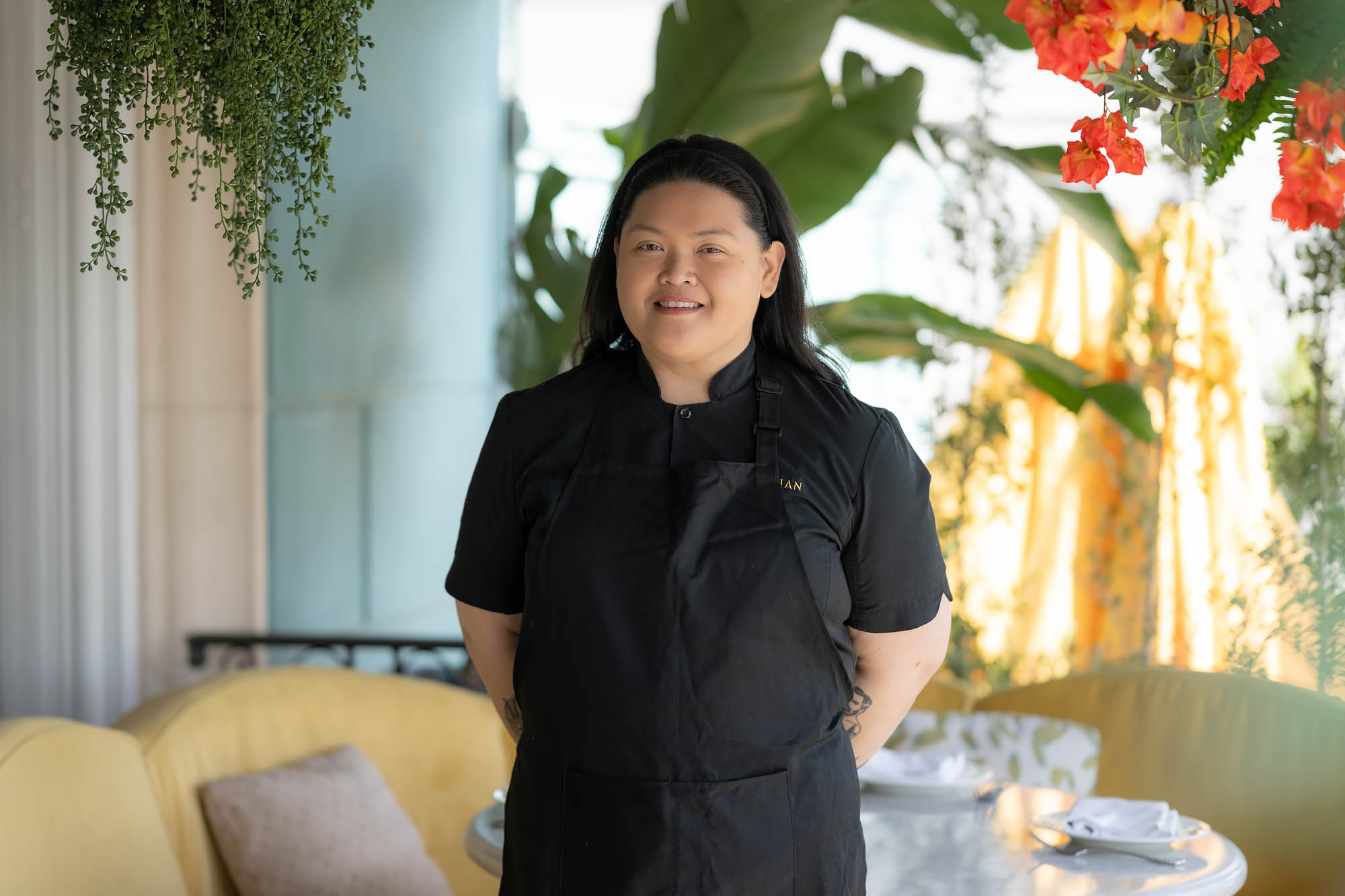A portrait of Thessa Diadem wearing a black apron and chefs coat while standing in the dining room at the Georgian hotel with flowers and greenery surrounding her. 