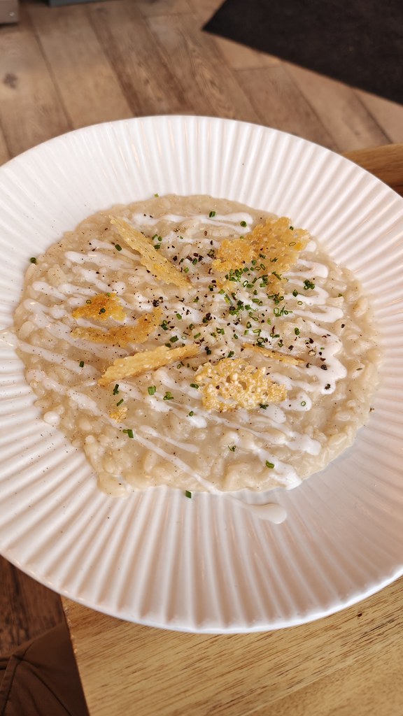 A large white bowl/plate type of dish with a large portion of risotto, drizzled with some sort of cream sauce and with chunks of baked parmesan and pepper on top.