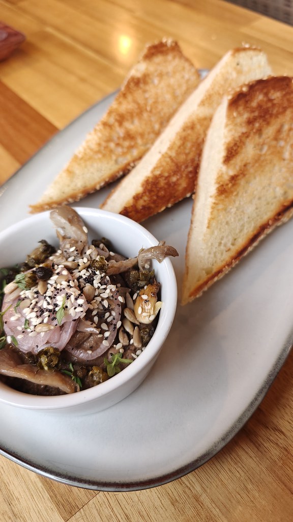 Three slices of toasted bread served alongside a small white bowl filled with the mushroom pate, which is topped with pickled shallot and sesame seeds.