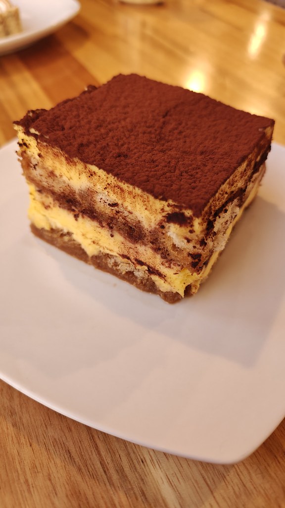 A small white plate with a big ol cube of tiramisu on it. It is a heck of a solid block of creamy white goodness and cocoa powder.