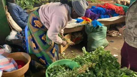 Vegetable vendor Priscilla Njeri - wearing a pale pink fleece, a green, blue and yellow print wrap as a skirt and grey beanie - bends as she cuts the roots off some vegetables with a knife at her market stall in Kiambu county