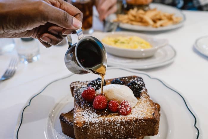A hand pours syrup over French toast topped with powdered sugar, raspberries, blackberries, and a dollop of cream, on a white plate