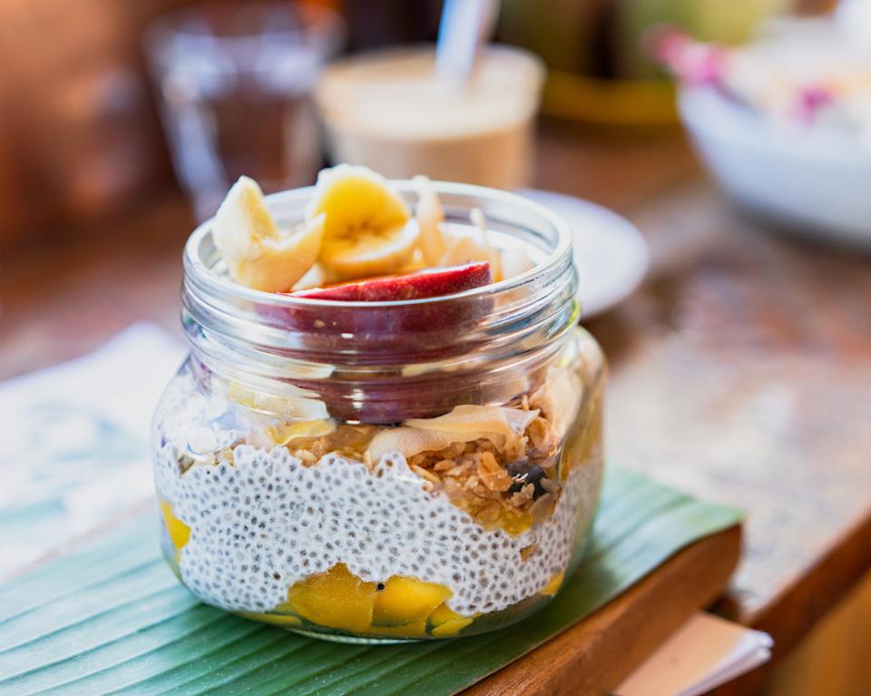 Jar of chia pudding with banana slices, apple, and granola on a banana leaf. Smoothie and salad blurred in the background