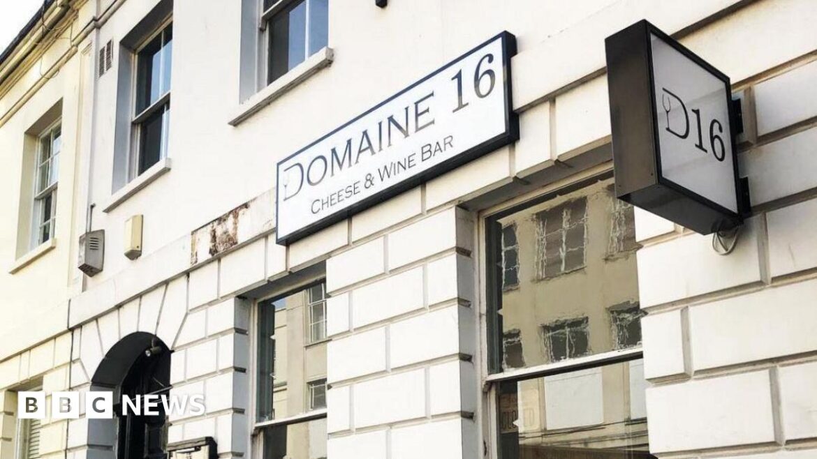 A white building with a number of windows and a black door. Two signs are on the wall, one reads Domaine 16 cheese and wine bar and the other says D16.