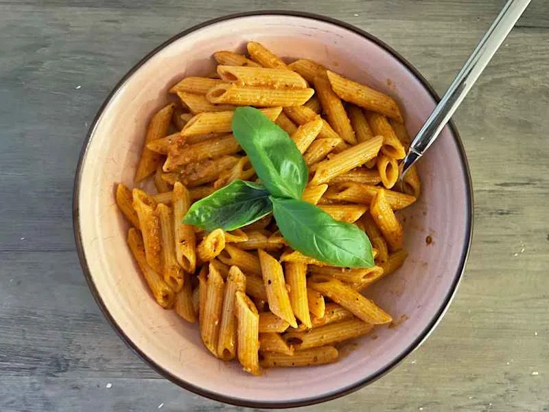 Penne pasta in a pink bowl topped with fresh basil leaves, served with a silver fork on a wooden surface.