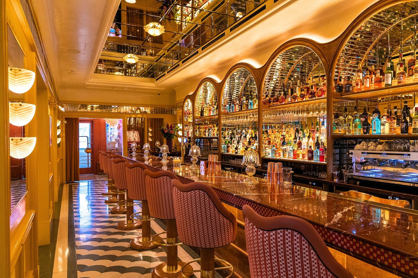 An impressive golden bar stretching down a corridor with mirrored arches filled with drinks, and bar stools upholstered in geometric pink patterns