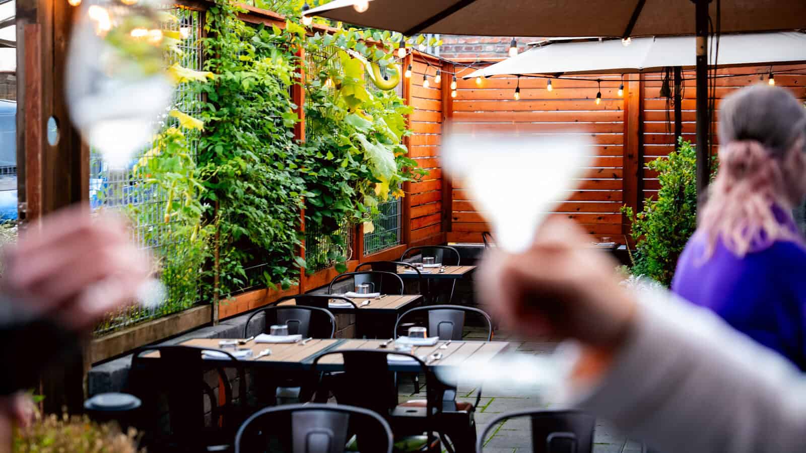 Outdoor patio with wooden fencing and greenery, empty tables and chairs, and people in the foreground holding up glasses for a toast—experience summer dining at its best when you visit Carrello.