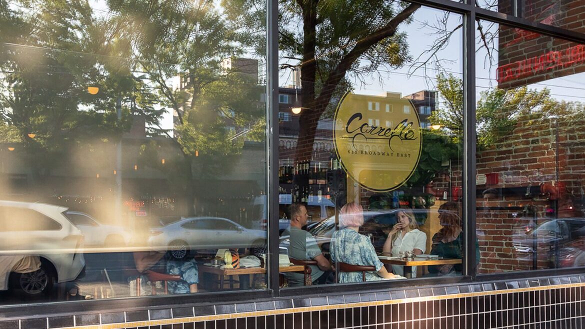 People are seated at tables inside Carrello café, seen through a large window with the café's name and street visible—a perfect spot for a summer visit. Cars and trees are reflected in the glass.
