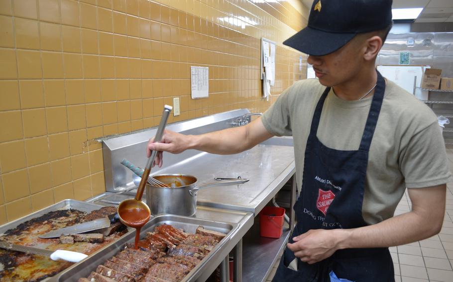 A man in a gray shirt, blue hat and blue apron uses a ladle to pour gravy onto a tray of meatloaf on a stainless steel counter in a kitchen.