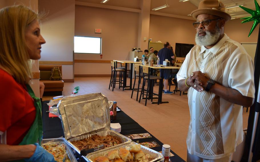 A man in a white, brown hat and glasses speaks with a woman in a red shirt and green apron with a table with trays of food between them.