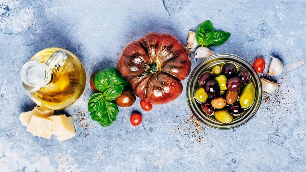 An aerial view of a bottle of olive oil, parmesan cheese, an organic tomato, basil leaves, a glass container of olives, and garlic, as staples of a Mediterranean diet