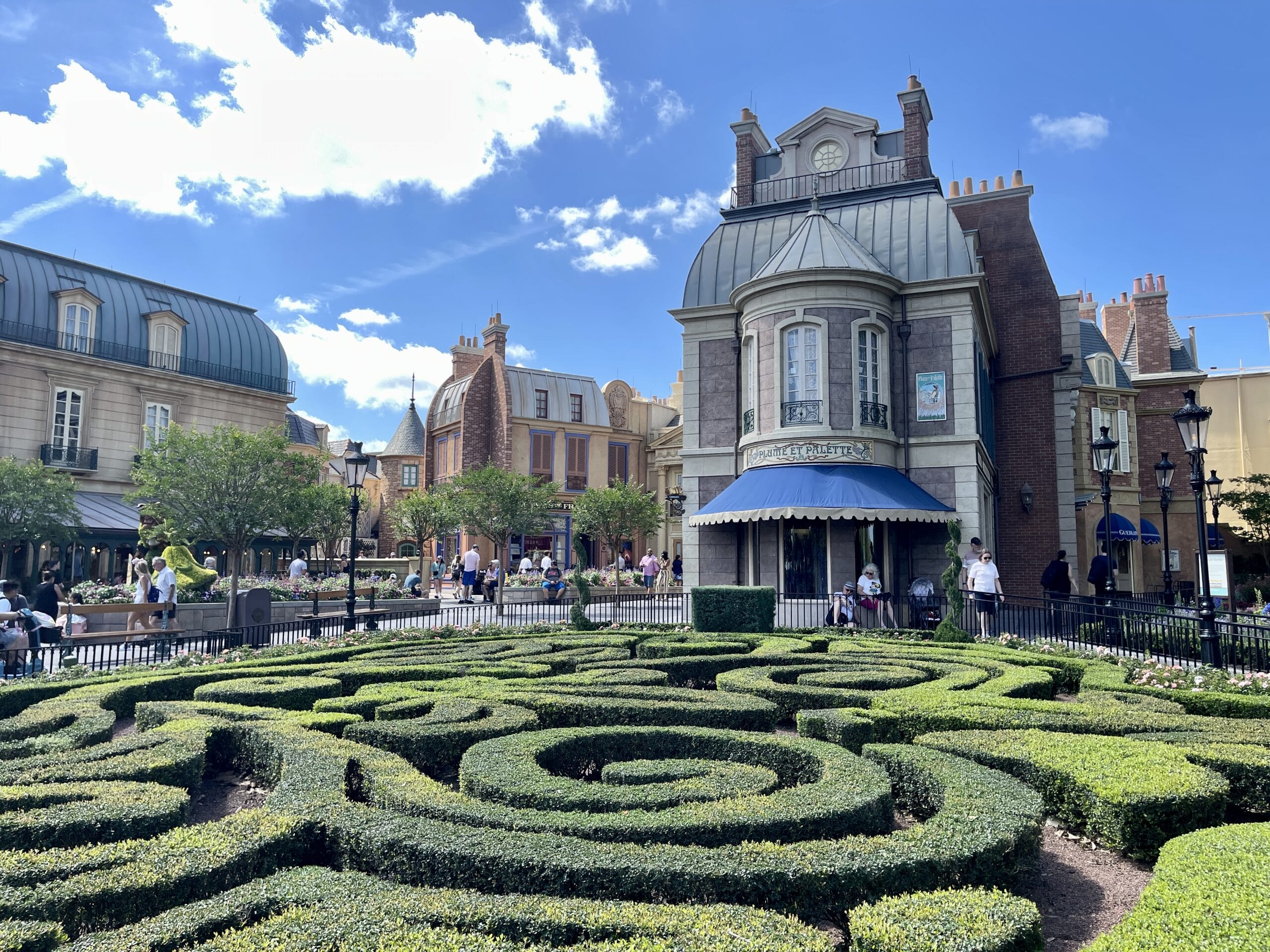 A neatly trimmed hedge maze is in the foreground, with a European-style building reminiscent of EPCOT in the background against a partly cloudy blue sky. Several people are walking and sitting around the area, enjoying the tranquil scene.