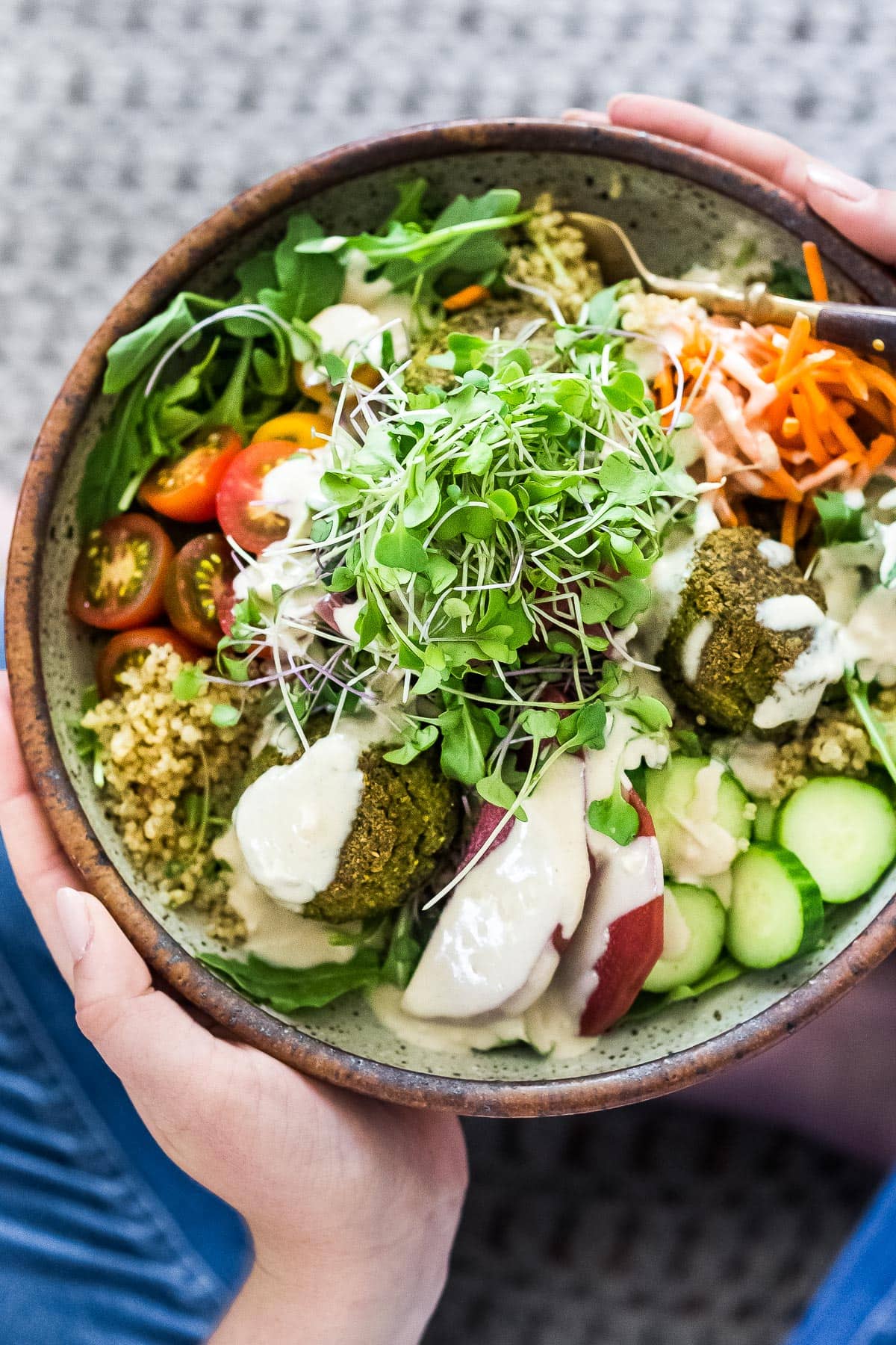 falafel bowl with sliced cucumbers, cherry tomatoes, shredded carrots, micro greens, grains, and drizzled tahini sauce.