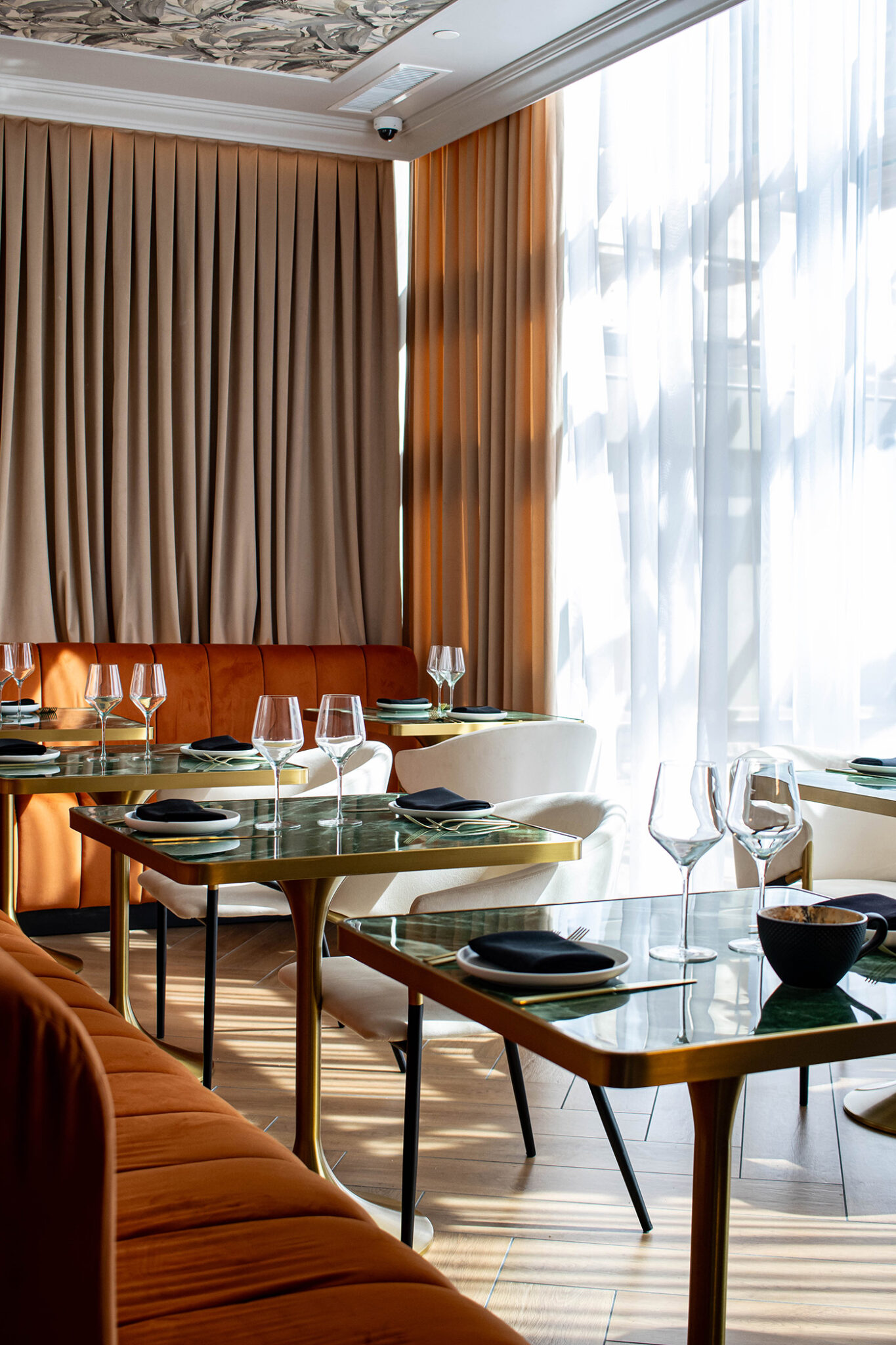 A restaurant dining room with tables and bench seating, glasses and cutlery on the tables, and full-height curtains over the windows.