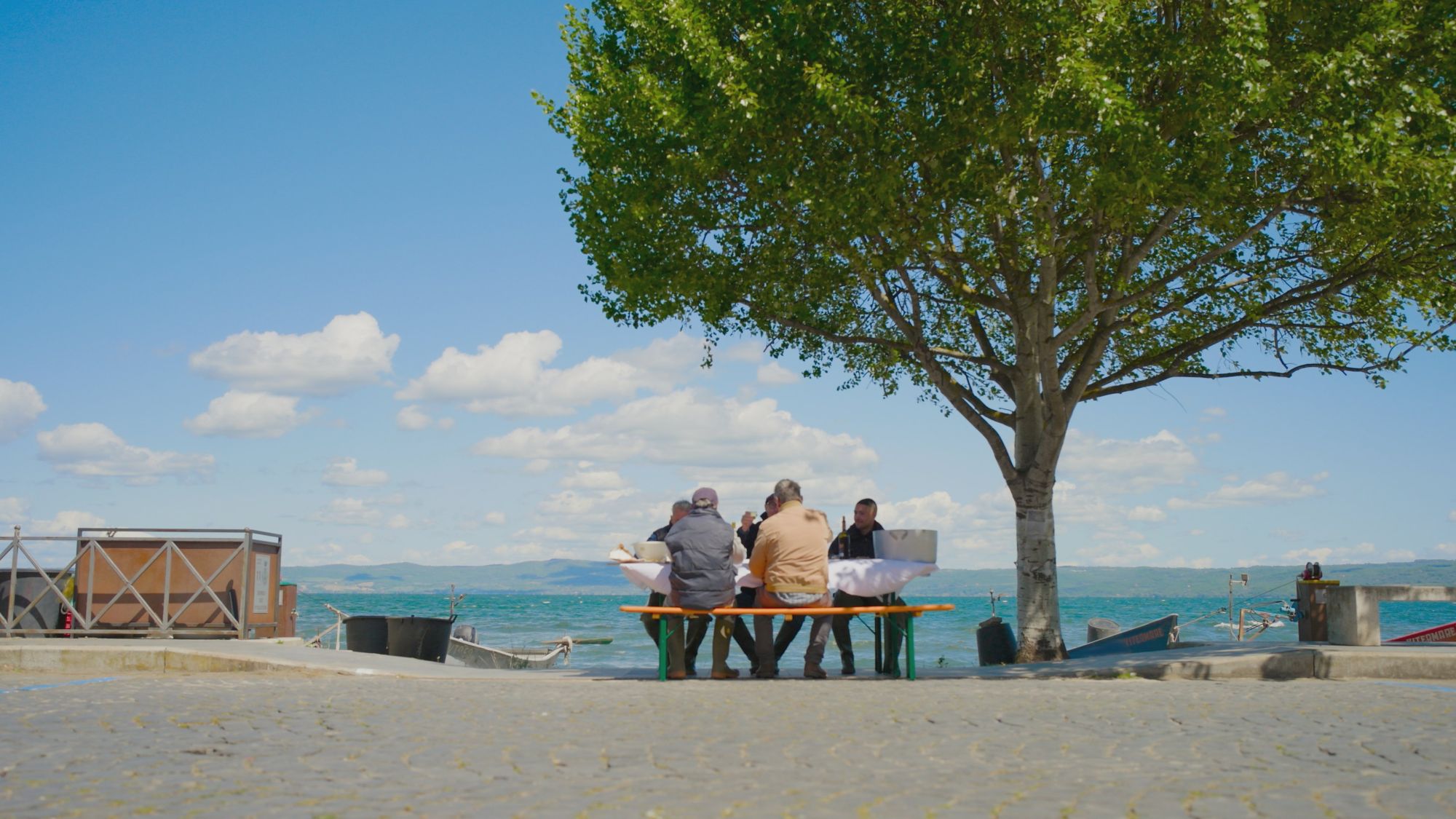 Stanley Tucci and local fisherman eat at a picnic table alongside a lake in Tucci in Italy
