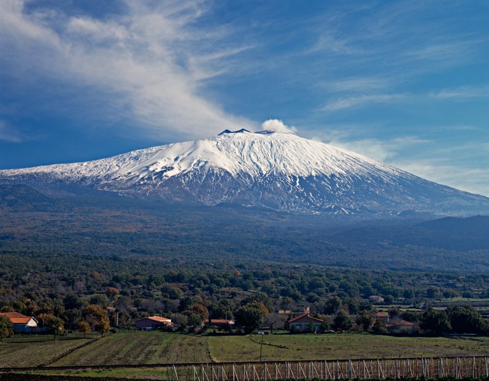 Mount Etna, snow-capped and smoking, overlooks Sicilian farms and vineyards.