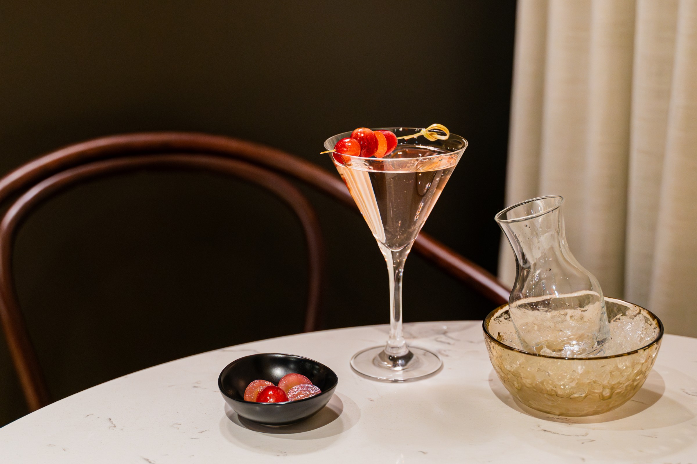 A pink-tinged martini sits on a table with a sidecar and a little dish holding the grape garnish.
