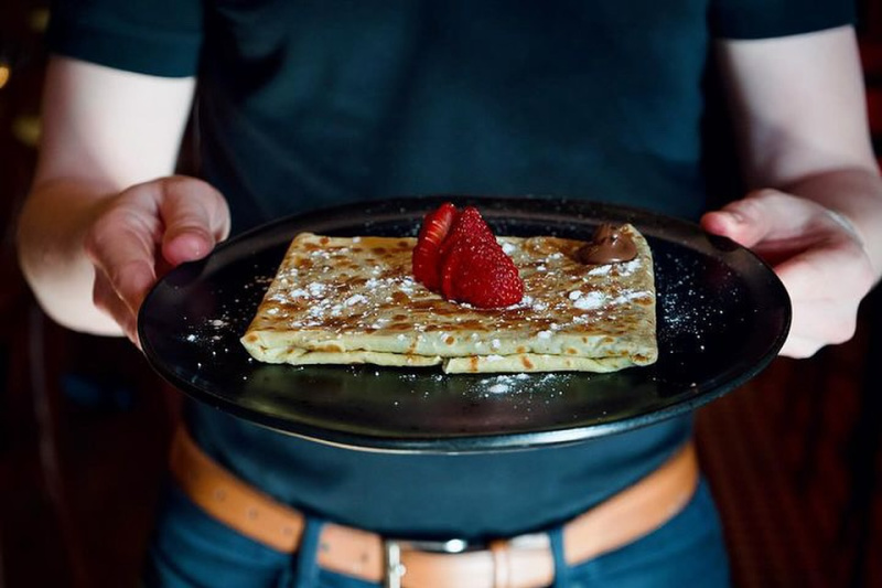 A person holding a black plate with a folded crepe topped with powdered sugar, sliced strawberries, and a dollop of chocolate spread—perfect for anyone craving delicious crepes in Nashville.