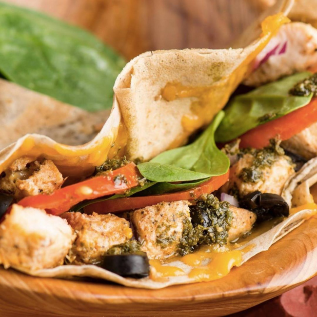 A close-up of a wrap filled with grilled chicken, spinach, tomato, olives, cheese, and pesto sauce on a wooden plate.