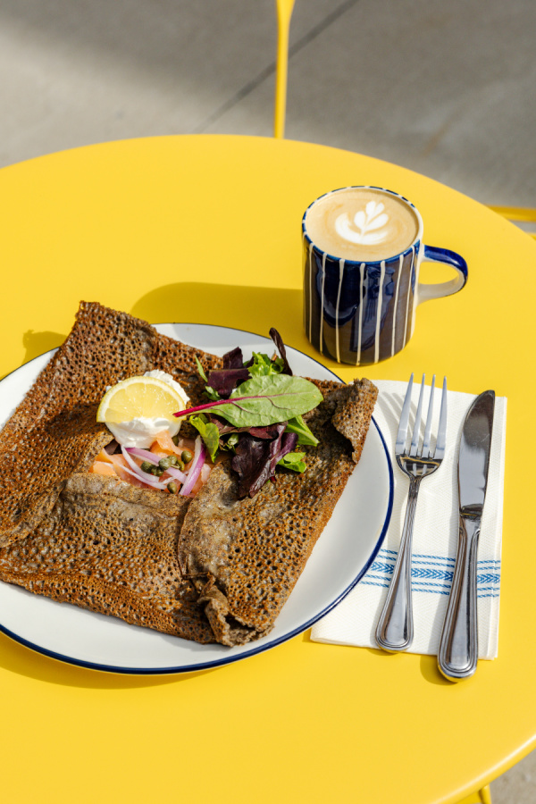 A plate with a buckwheat crepe, salad, and lemon wedge sits on a yellow table next to a cup of latte with latte art—perfect for enjoying crepes in Nashville with a fork and knife on a napkin.
