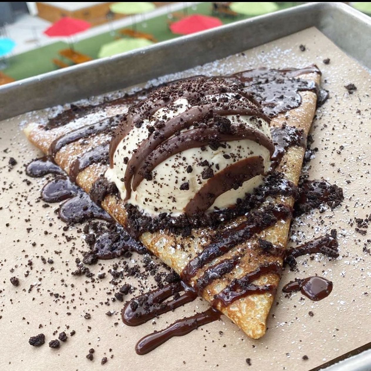 A French Crêpe topped with a scoop of vanilla ice cream, chocolate drizzle, crushed cookies, and powdered sugar on a parchment-lined tray.