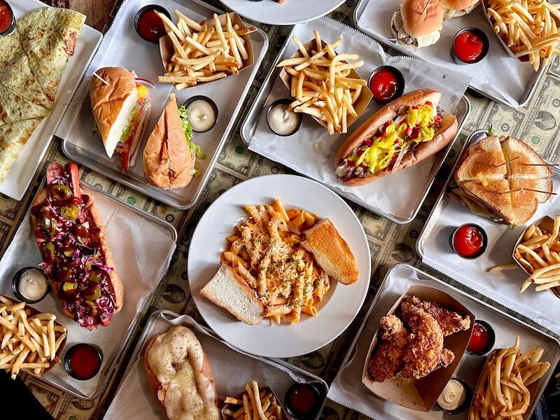An overhead view reveals a festive spread from Memphis restaurants open on Christmas, featuring an array of dishes like sandwiches, fries, pasta, hot dogs, and fried chicken, all served on trays with ketchup and mayonnaise.