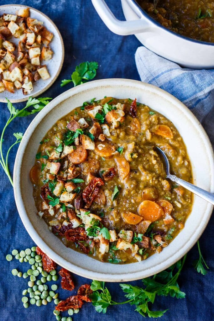 Mediterranean split pea soup in bowl with spoon, topped with croutons, sun-dried tomatoes, and fresh herbs. 