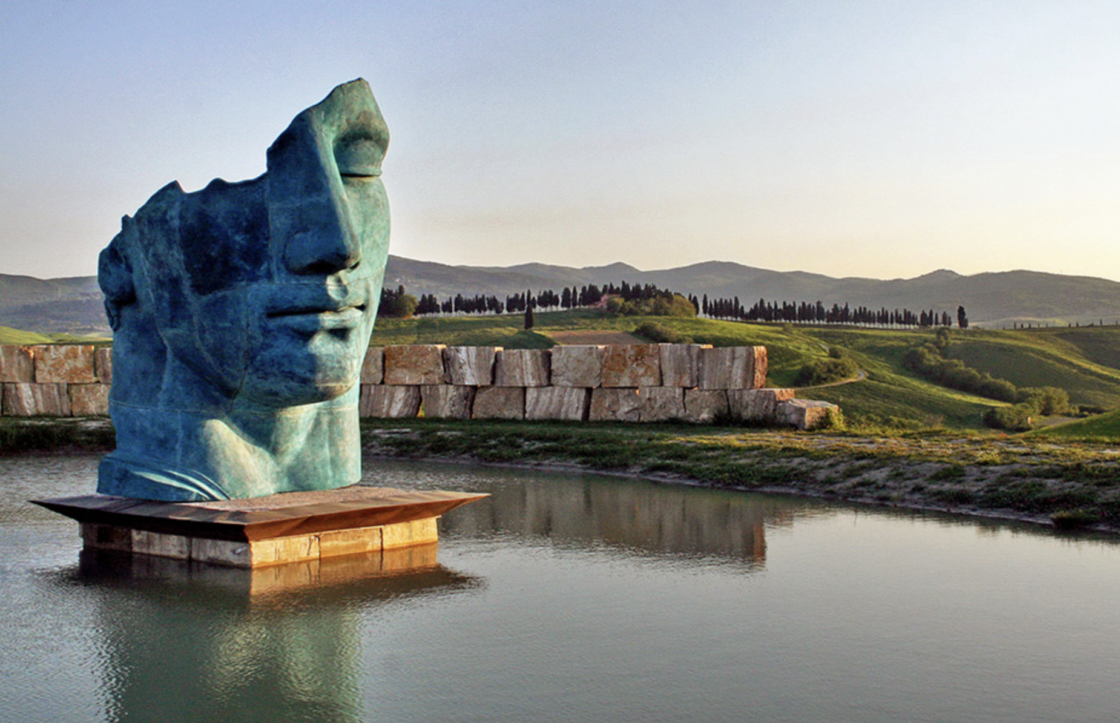 Image of a larger-than-life sculpture of a Roman head floating on a lake, surrounded by vineyards.