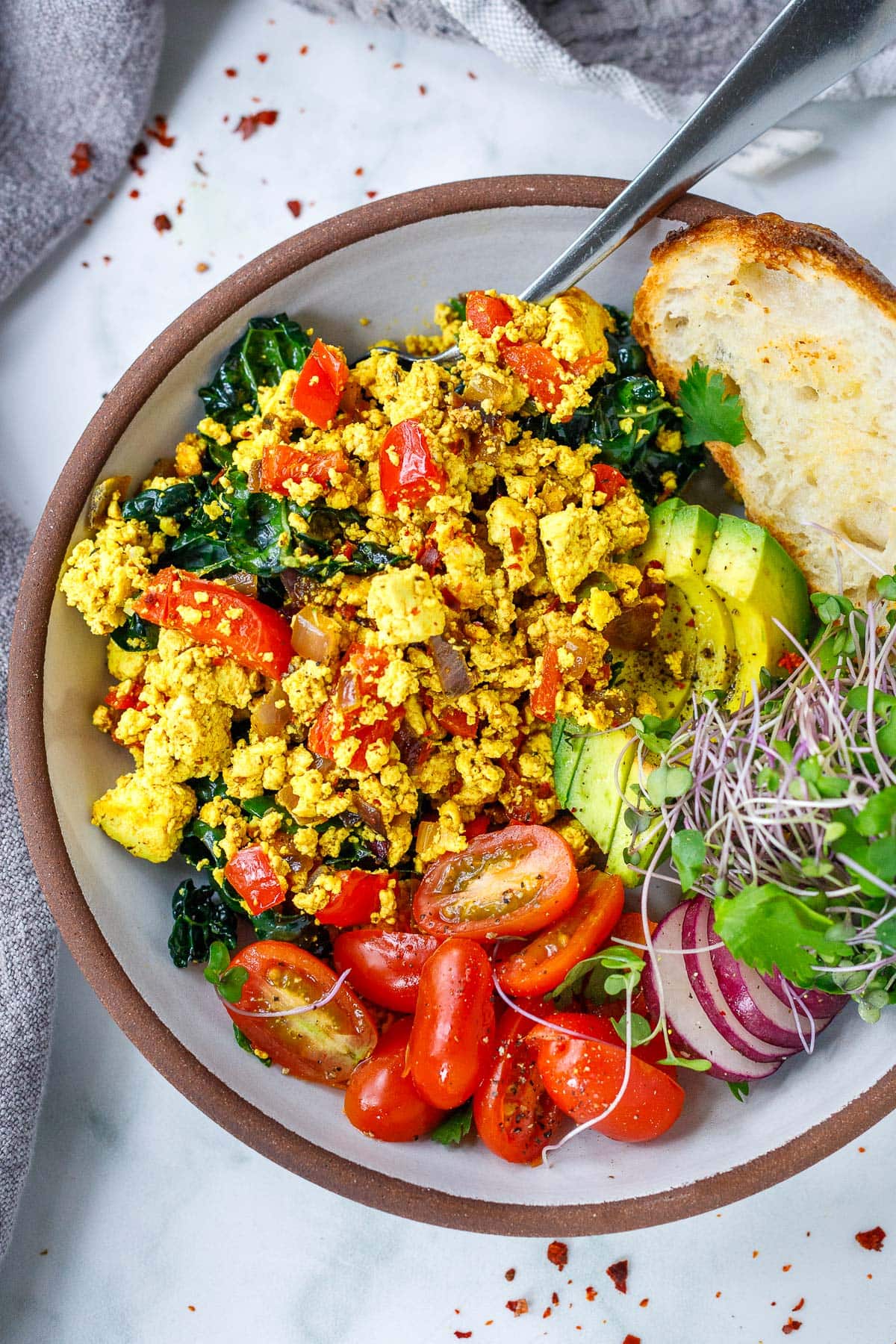 tofu scramble with greens and veggies, served with sliced avocado, tomato, radish, micro greens, and toast.