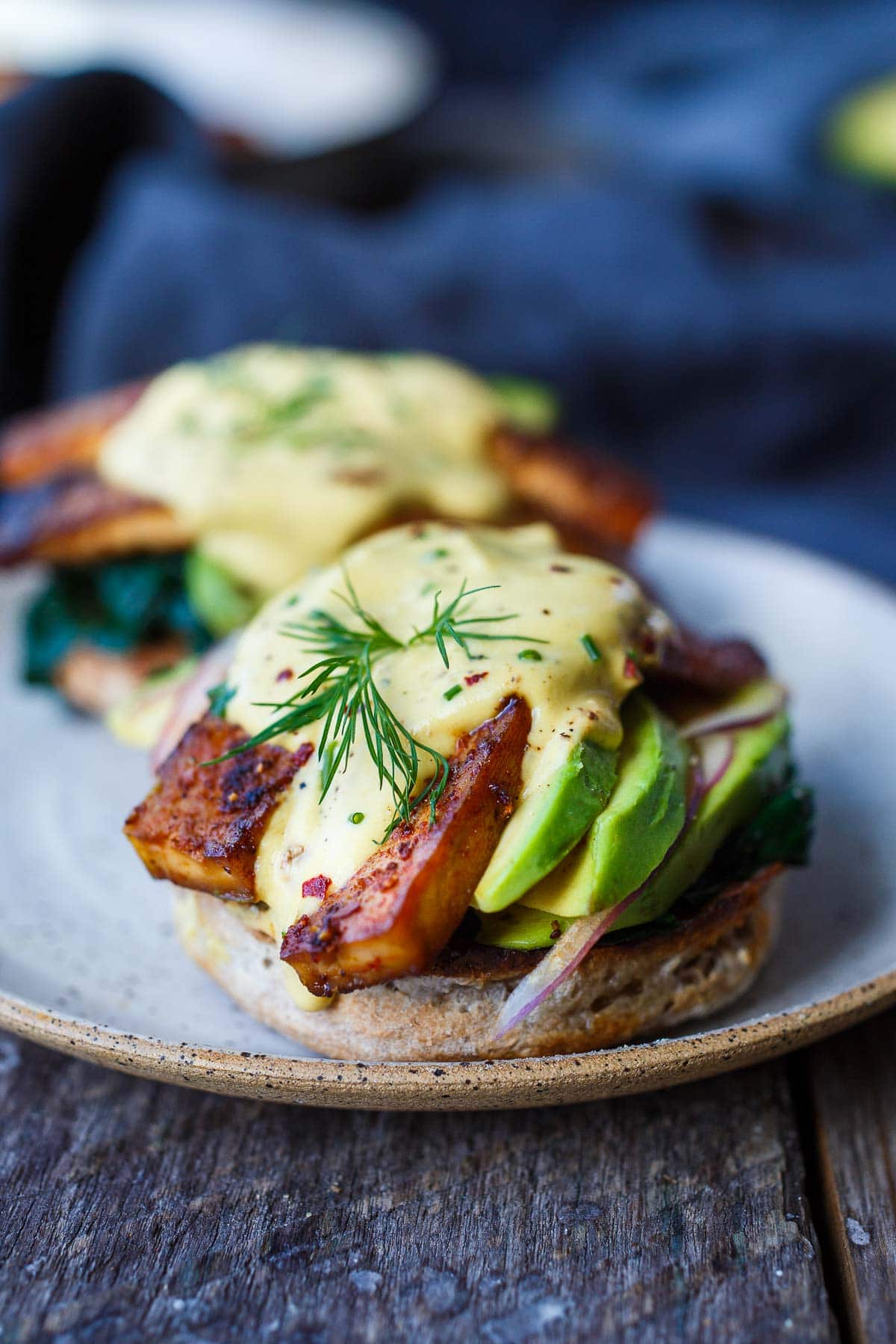 opened faced english muffin with vegan eggs benedict, made with tofu bacon, sliced avocado, sliced red onion, topped with vegan hollandaise and fresh dill.