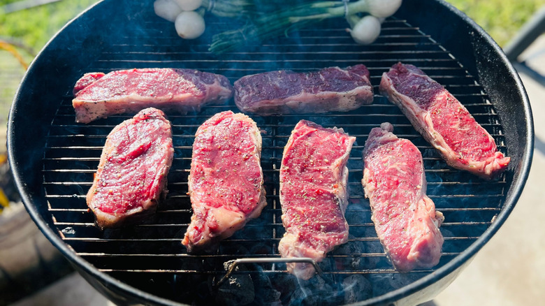 Seven raw NY strip steaks cooking on black grill