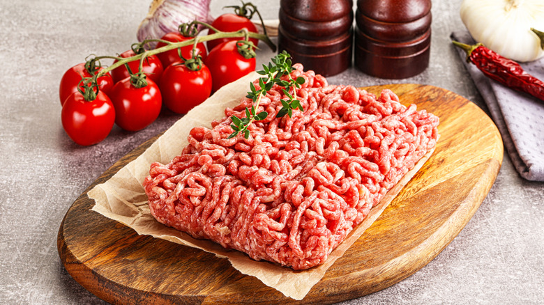Raw ground beef on a wooden board topped with rosemary with tomatoes and pepper mills in the background