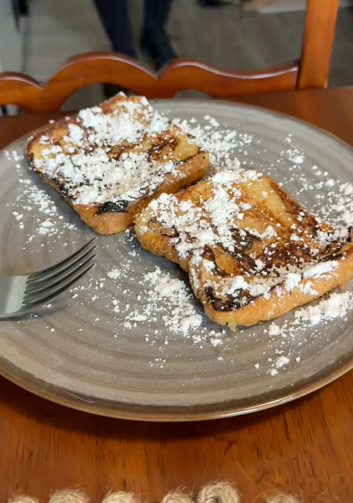 French toast topped with powdered sugar on a plate with a fork. A blurred background shows a person and a resting dog