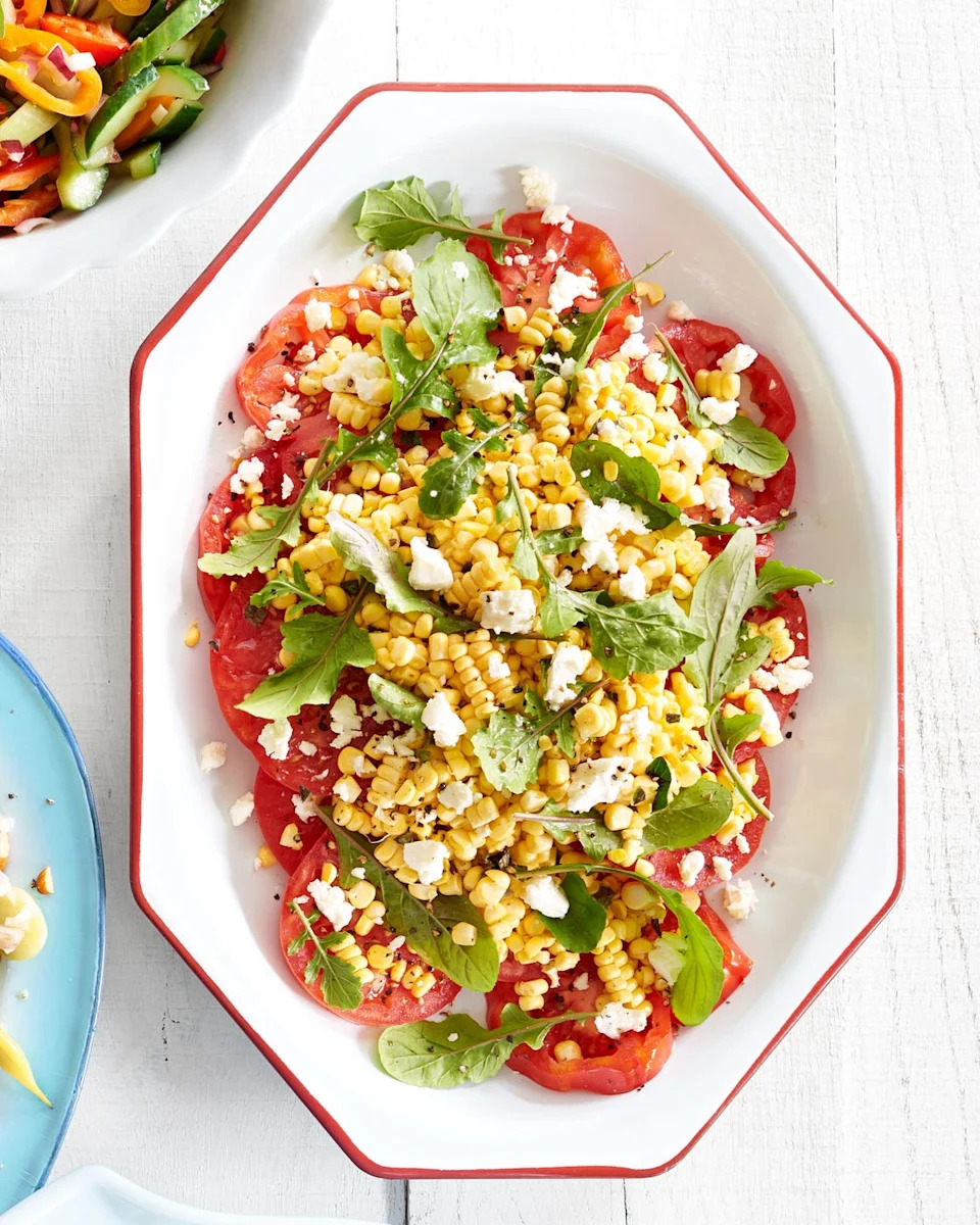 sliced tomatoes topped with corn, greens, and crumbled cheese on a platter
