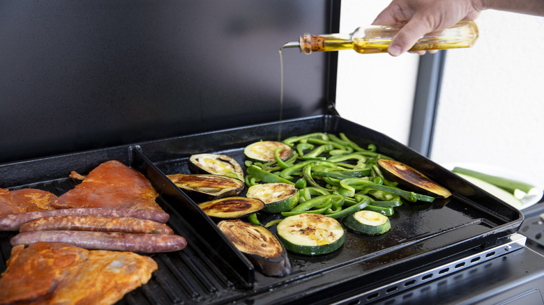 Close up of a person pouring olive oil on a plancha grill with meat and vegetables