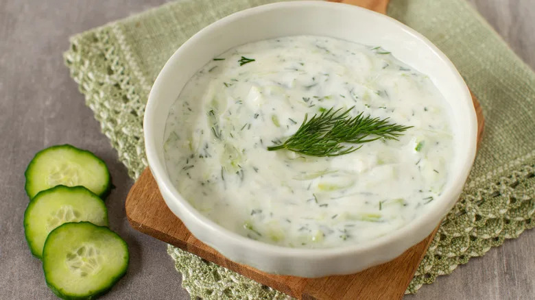 White bowl of tzatziki sauce garnished with fresh dill on a wooden board with a green kitchen cloth and some cucumber slices on the side