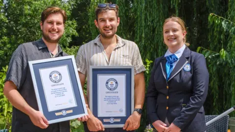 Oli Paterson and Phoenix Ross Phoenix, on the left, is stood smiling holding a Guiness World Record certificate, next to Oli, in the middle, who is also smiling and holding a certificate. The female adjudicator, in a blue uniform is stood on the right also smiling. They are outside, with large green trees behind them. 
