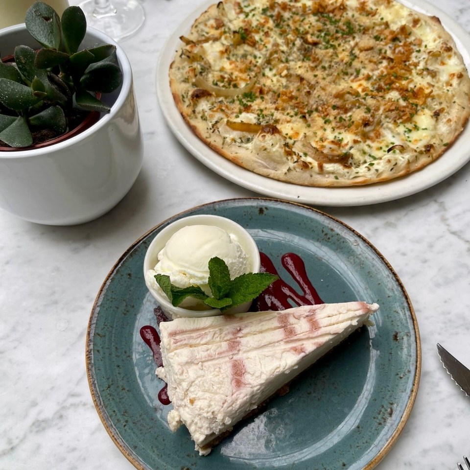Cheesecake slice with ice cream and a pizza in the background.