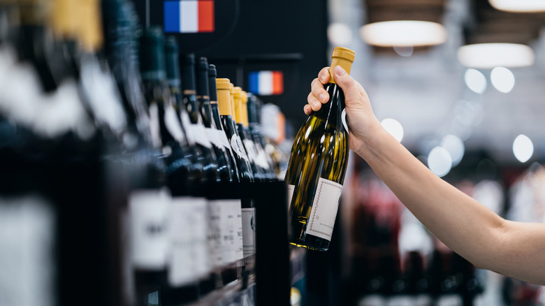 A hand picking out a bottle of wine in the French wine aisle, with a bokeh background