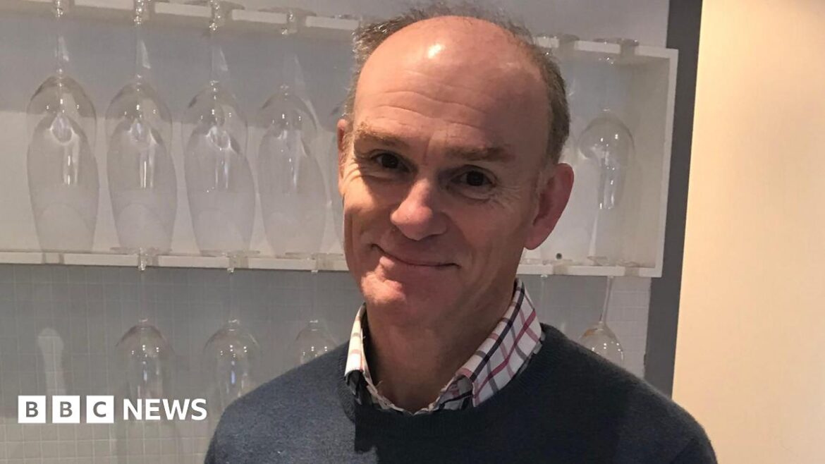 Philip Tuck in a blue jumper with a checked shirt, standing in front of shelves with wine glasses on.